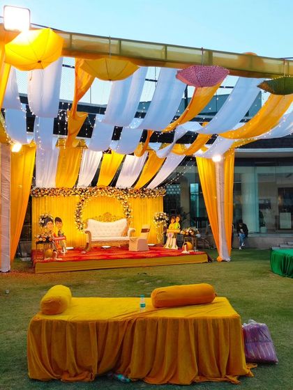 An outdoor Haldi event with a large canopy of yellow and white drapes. The setup includes a decorated stage and informal floor seating for a relaxed vibe.
