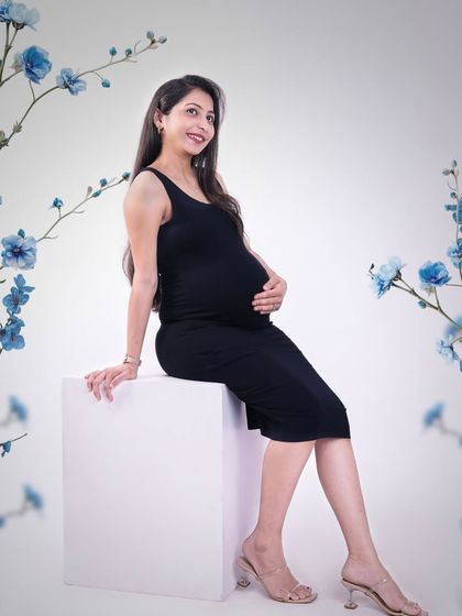 A simple and elegant studio portrait. The mother-to-be is dressed in a classic black dress against a clean white background, framed by delicate blue flowers for a pop of color.