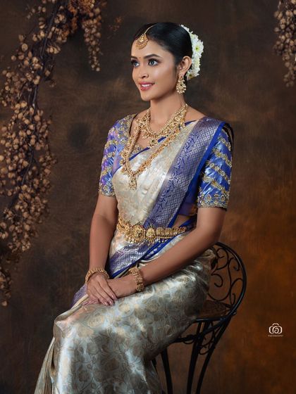 A seated portrait showcasing a beautiful silver and blue silk saree. The warm, earthy backdrop is decorated with floral elements, adding a soft, romantic touch to the traditional attire.