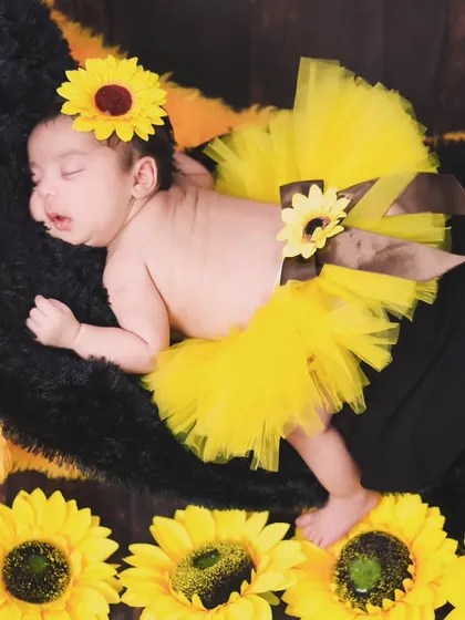 A full-body shot of a newborn in a sunflower-themed tutu, sleeping amongst bright yellow flowers.