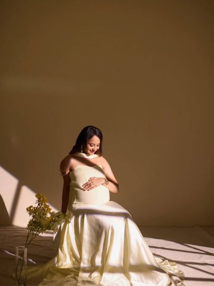 A minimalist setup with beautiful, soft light. The simple play of light and shadow across her gown creates a serene and artistic motherhood portrait, focusing on the gentle curve of her bump and her peaceful expression.