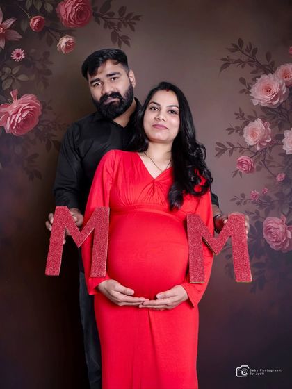 A classic couple's maternity portrait against a floral backdrop. The mom-to-be is in a vibrant red dress, holding 'MOM' letters with her partner.