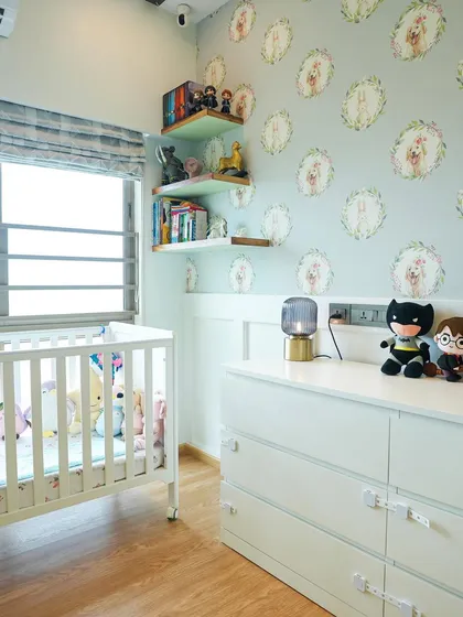 The nursery features a white crib and dresser, floating shelves for books and toys, and a soft, patterned Roman blind.