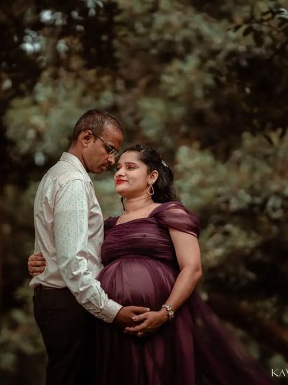 A classic couples maternity portrait set against a lush, green background. The soft, natural lighting emphasizes their gentle embrace and the beautiful texture of her deep purple gown.