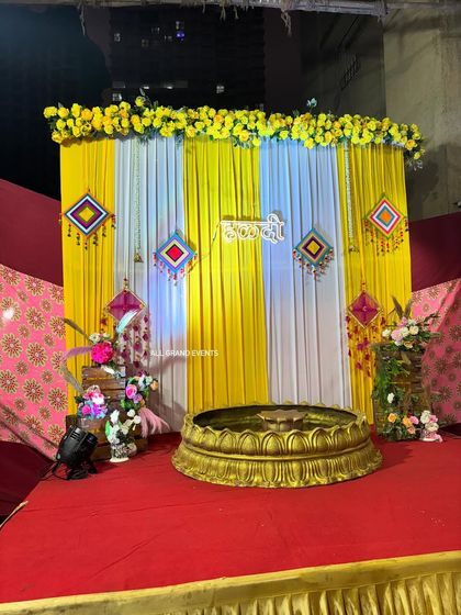 A full view of the Haldi stage setup at night. The lighting highlights the bright yellow drapes and the intricate details of the traditional hangings.