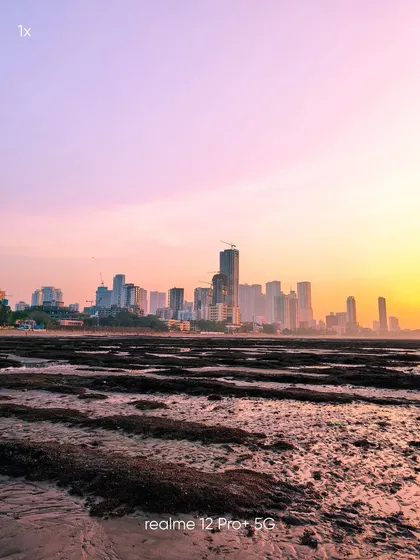 A wide-angle view of the Mumbai skyline at sunset, taken from the shoreline. This mobile photograph captures the expansive cityscape and the beautiful colors of the sky reflecting on the wet sand.