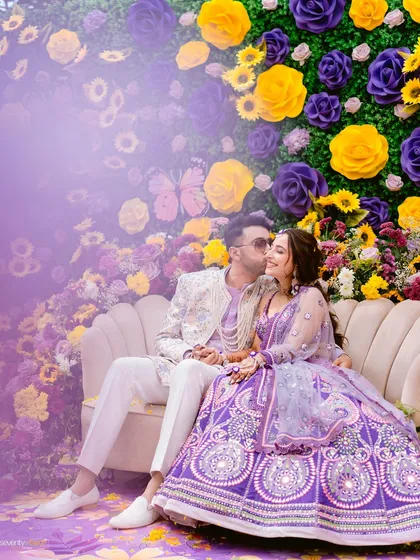 The couple shares a tender moment on a couch surrounded by a wall of purple and yellow flowers, a beautiful and romantic portrait from their Haldi.