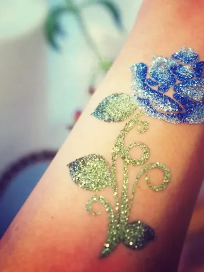 A close-up of a beautiful rose tattoo made with blue and silver glitter on a green stem. The glitter gives it a frosted, magical appearance.