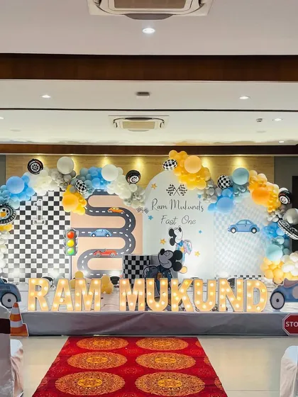A 'Two Fast Two Curious' car-themed first birthday party. This large-scale setup features a custom printed backdrop with a racetrack, a multi-color balloon arch, car cutouts, and light-up marquee letters spelling out the birthday boy's name.