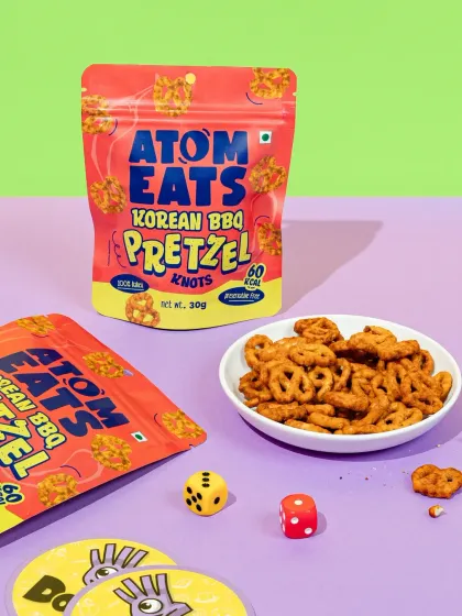 Atom Eats Korean BBQ Pretzels styled with game pieces against a colorful background. This playful setup suggests the snack is a perfect companion for fun and games.