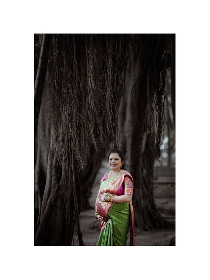 An atmospheric portrait of an expecting mother in a vibrant green and pink saree, standing before a large banyan tree. This photo from her baby shower shoot has a timeless, natural feel.