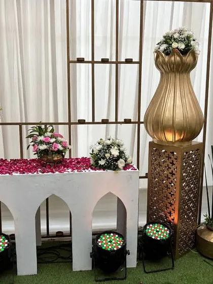 A traditional decor setup on a white arched table, featuring golden vases, flower petals, and intricate pedestals, creating an elegant and sacred corner.