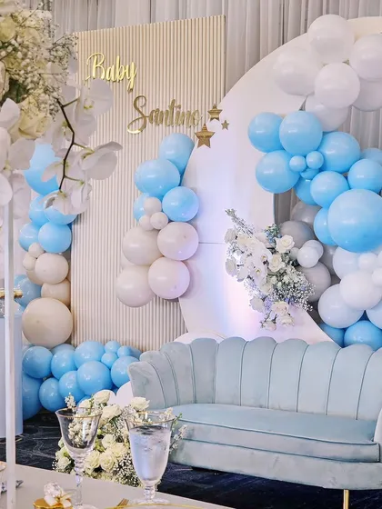 A serene and beautiful baby shower decor in shades of baby blue and white. The setup includes a crescent moon backdrop, a comfortable couch for the mom-to-be, and elegant white florals.