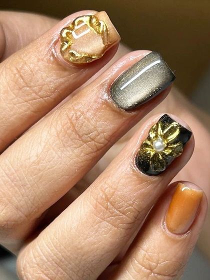 A close-up of a luxurious 3D nail art design. This look features a cat-eye base with sculpted gold and black floral elements, accented with a single pearl.