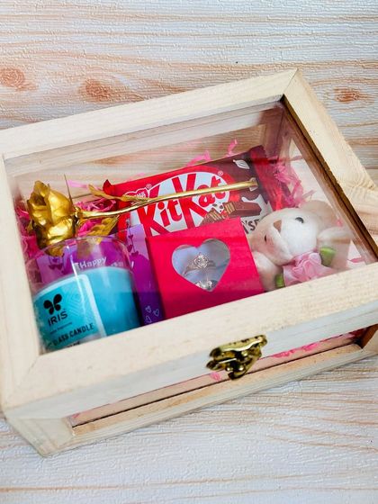 A mini Valentine's hamper with a golden rose. This wooden box includes a scented candle, couple rings, a teddy bear, and chocolate for a sweet and romantic surprise.