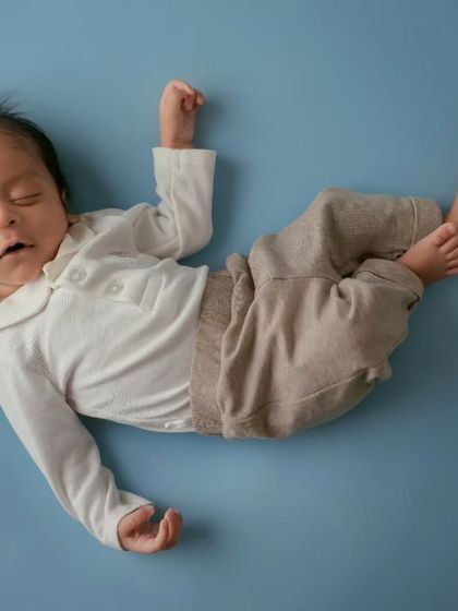 A newborn in a white polo shirt and tan pants sleeps peacefully on a solid light blue background, creating a clean and classic look.