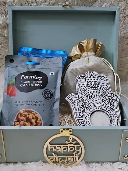 This premium trunk box features a "Happy Diwali" cutout and is filled with healthy snacks like black pepper cashews and salted almonds, along with a beautiful Hamsa tealight holder.