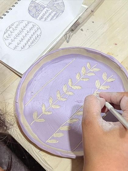 A student uses their sketchbook for inspiration while painting a delicate leaf pattern onto a plate. We encourage you to bring your own designs to class.