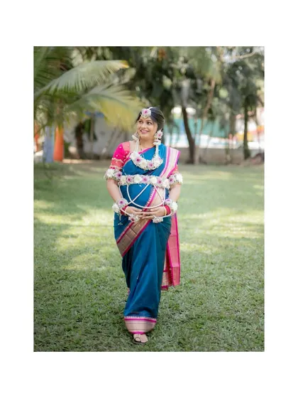 A full-length portrait of the radiant mother-to-be, showcasing her traditional blue and pink saree and the beautiful floral ornaments for her baby shower.