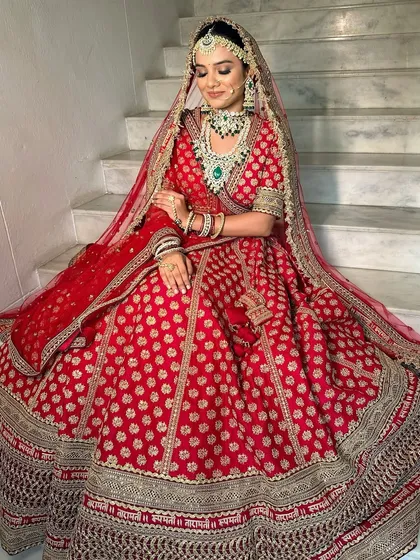 A full-length shot of the bride seated on the stairs, showcasing the grandeur of her red lehenga and the elegance of the complete styling.