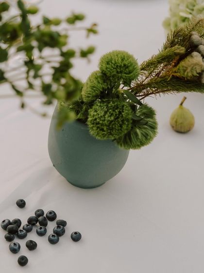 A close-up of a small green vase with green pom-pom flowers and scattered blueberries, part of the organic tablescape.