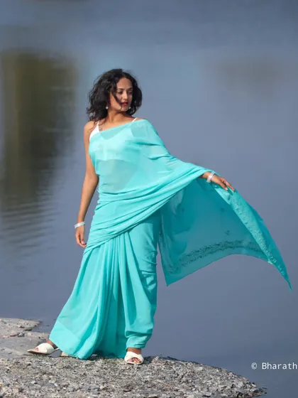 A full-length portrait by the water, showcasing the vibrant color of the turquoise saree. The model's pose is strong and confident.