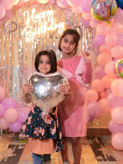 Two friends holding a heart-shaped balloon, posing in front of a pink balloon wall.