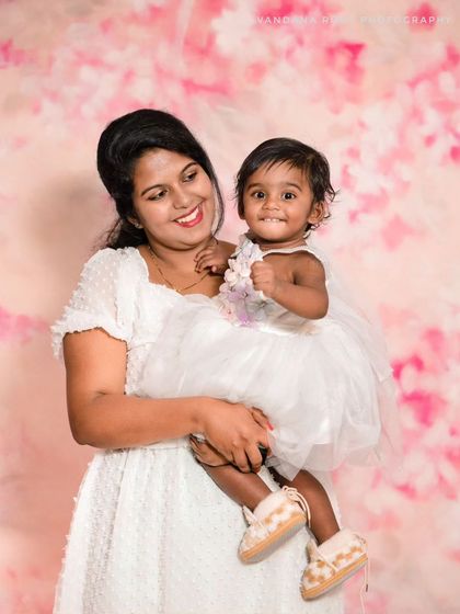 A mother and her smiling daughter pose against the same pink floral backdrop. Their matching white dresses and happy expressions make for a beautiful Mom & Me portrait.