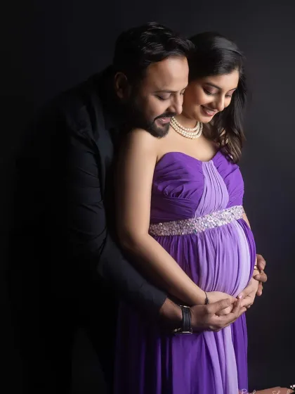 An intimate and tender moment between expectant parents. Posed against a classic dark studio backdrop, this portrait focuses on their close bond and shared excitement, with his arms wrapped lovingly around her and the baby bump.