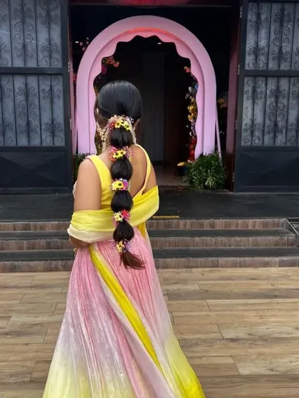 A full-length back view of a bride with a floral bubble braid, complementing her colorful ombre lehenga. It's a perfect hairstyle for a playful and festive Mehendi party.