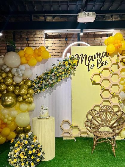 A vibrant 'What Will It Bee' baby shower. We used a honeycomb-patterned backdrop, a wicker peacock chair, and a cheerful yellow, white, and gold balloon garland to create this buzz-worthy celebration.