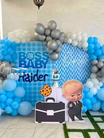 This is a clean and modern Boss Baby setup for Haider's first birthday. It features a custom checkered backdrop, a large character cutout with a prop briefcase, and an organic balloon garland in blue, white, and silver.