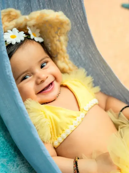A close-up shot capturing a radiant smile. The daisy headband and frilly yellow outfit add to the sunny, cheerful feel of this beach-themed infant photography session.