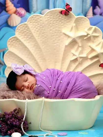 A tiny newborn mermaid, wrapped in purple, sleeps soundly in a clamshell cradle in our magical underwater world.