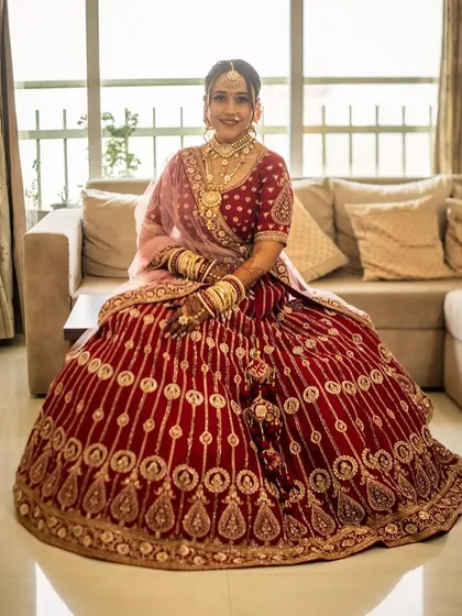 A full-length portrait of the bride seated, showcasing her magnificent red velvet lehenga. The makeup is designed to complement the richness of the fabric and the intricacy of the embroidery.