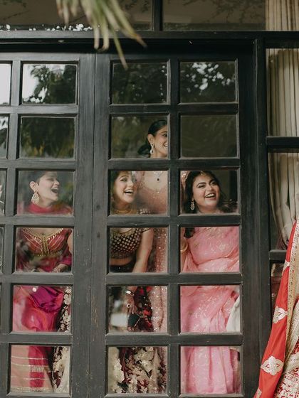 A fun and candid moment of the bride Ashmeet with her bridesmaids peeking through the window panes. It's a perfect capture of the excitement and friendship on the wedding morning.