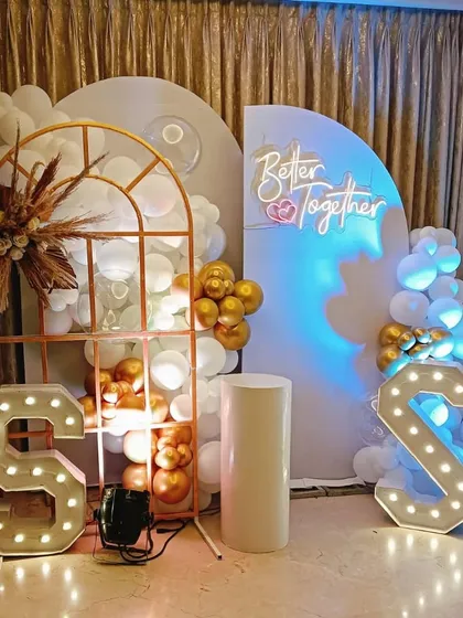 A chic "Better Together" anniversary setup with marquee initials, a neon sign, and a modern arched backdrop decorated with pampas grass and neutral-toned balloons.