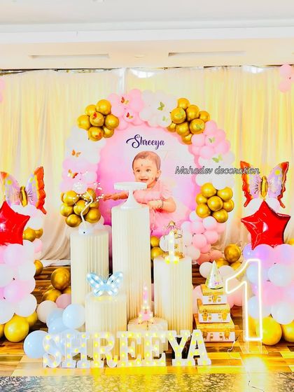 A personalized first birthday party with a butterfly theme. The round backdrop includes a photo of the birthday girl, framed by a pink and gold balloon garland and accented with butterfly and star foil balloons.