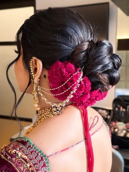 A voluminous and intricately textured bridal bun, perfect for a North Indian wedding ceremony. This style is accented with deep magenta flowers and a delicate chained accessory, creating a look that is both classic and striking from every angle.