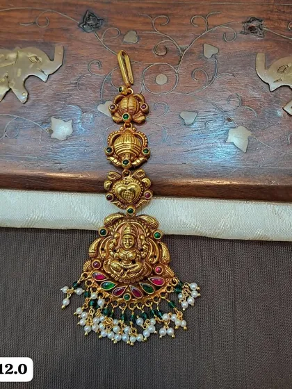This lightweight maang tikka is made from hallmarked 92.5 silver with gold plating. It features a Goddess Lakshmi motif and is adorned with colorful stones and pearl drops.