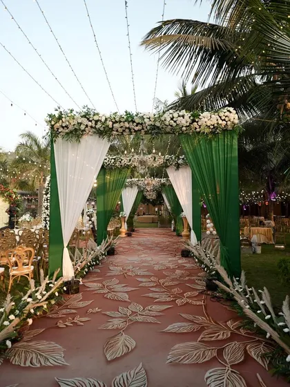 A stunning entrance walkway for a wedding ceremony. The combination of green and white drapes, a floral-printed carpet, and elegant chandeliers creates a grand and welcoming path for guests.