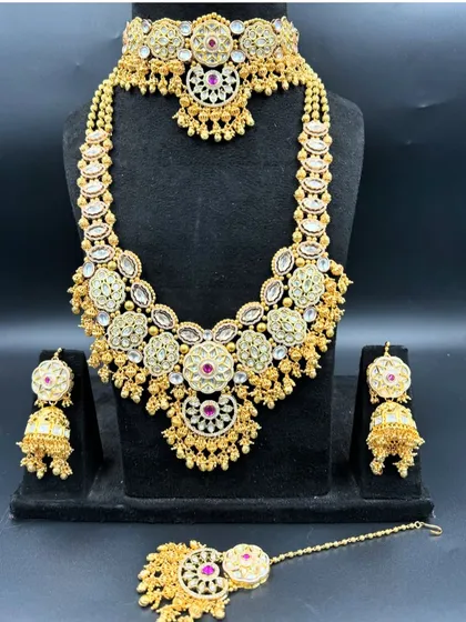 This bridal rental set features beautiful ruby-coloured stones set in an intricate gold-tone design. It includes a choker, a matching long haar, jhumkas, and a maang tikka to complete your wedding ensemble.