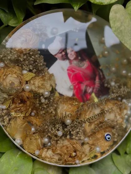 A circular keepsake with preserved white flowers and pearls, set against a backdrop of green leaves. The couple's photo is perfectly centered in this beautiful arrangement.