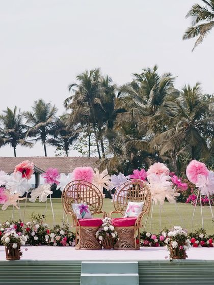The main stage for a pastel beachside Mehendi in Goa, featuring whimsical paper flowers and comfortable wicker seating, all set against a backdrop of palm trees.