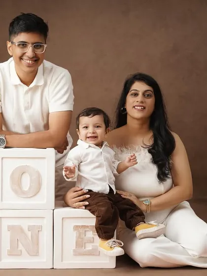A warm family portrait with a brown backdrop, showing two mothers and their son celebrating his first birthday with 'ONE' blocks.