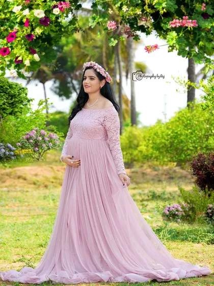 A serene portrait of a mother-to-be in a pink lace gown and floral crown, standing in a beautiful garden setting.