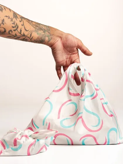A tattooed arm holds a patterned reusable bag, demonstrating its use. The clean white background and simple action make this an effective and relatable e-commerce shot that shows scale and function.