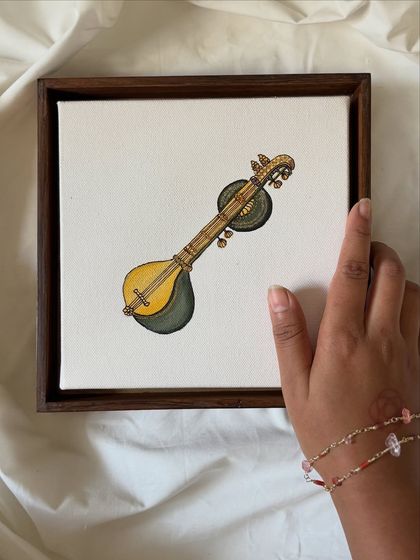 My hand holding a miniature painting of a Saraswati veena. This shows the small scale and intricate detail of the artwork, which is designed to be a precious, personal object.