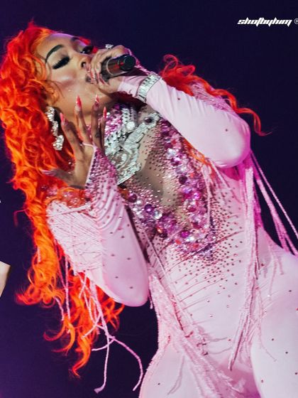 A female performer with vibrant orange hair sings passionately during Akon's Bangalore concert. The shot is filled with color and energy.
