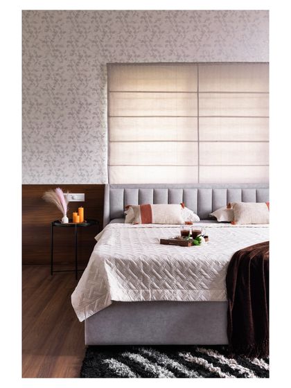 This cozy bedroom features a mix of textures for a warm and inviting feel. A soft upholstered headboard is set against a backdrop of subtle wallpaper and Roman blinds, creating a layered and comfortable space.
